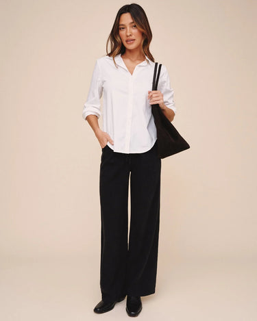 A woman modeling a white long-sleeve button-down shirt and black wide-leg trousers, carrying a black shoulder bag against a solid beige studio background.
