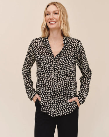 A woman modeling a black long-sleeve button-up blouse with a small white floral or ditsy print, paired with black pants, against a solid beige background.