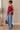 Woman wearing a red sleeveless top and blue jeans, standing indoors with a neutral background.