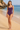 A woman modeling a purple ruched one-piece swimsuit on a sandy beach with ocean waves and rocks in the background.