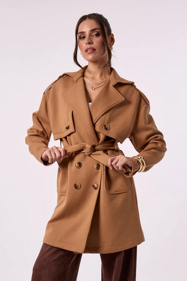 A woman wearing a camel brown belted trench coat with shoulder epaulets, paired with brown trousers, against a plain light background