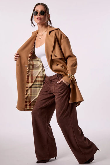 A woman wearing wearing a camel brown belted trench coat with shoulder epaulets, opening the coat to show the plaid lined interior paired with brown trousers, against a plain light background