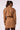 A woman shown from the back wearing a camel brown belted trench coat with shoulder epaulets, paired with brown trousers, against a plain light background.