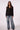 A woman modeling a black blazer over a black top with wide-leg blue jeans, standing against a light gray studio background.
