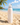 Pink Palm patterned water bottle with a silver cap on a sandy beach with ocean and sky in the background.