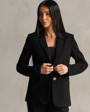 a woman wearing a black blazer with gold button accents and black pants with a white stripe on a neutral background