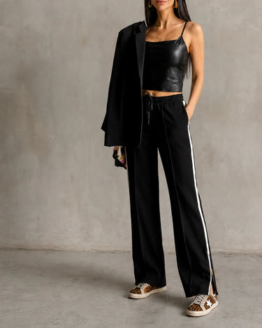 A woman wearing a black blazer and black pants with a white stripe and a zipper detailing at the hem on a neutral grey background.