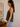Woman wearing a dark tank top and light blue jeans standing in a room with a wooden shelf and framed abstract art.