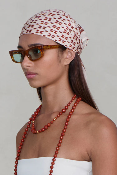 Woman wearing a patterned headscarf, sunglasses, and a long red necklace against a plain background