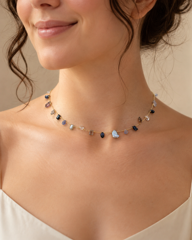 A close-up of a woman wearing a delicate multi-colored gemstone choker necklace with a gold chain, set against a soft beige background.