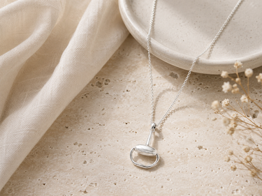 A silver pendant necklace with a minimalist ring and teardrop design, displayed on a textured beige stone surface next to a cream linen cloth and dried flowers.