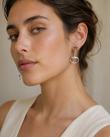 A close-up of a woman wearing a silver drop earring with a rectangular clear stone and an oval dangling element, paired with a cream-colored top against a neutral beige background.