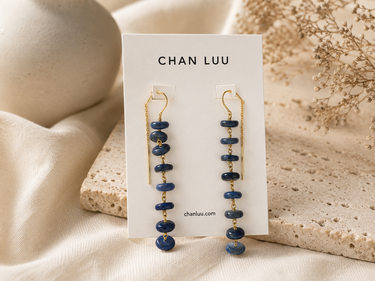 A pair of Chan Luu dangle earrings with dark blue stone beads and gold-toned metal, displayed on a white brand card against a warm beige fabric and stone background.