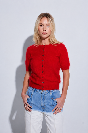 A blonde woman modeling a short-sleeve red knit cardigan with front buttons, paired with light blue jeans and white pants, standing against a light gray studio background.