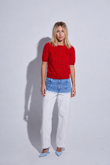 A woman stands against a light gray studio background wearing a bright red short-sleeve button-front top, a color-block denim skirt with blue on top and white below, white pants, and light blue pointed shoes.