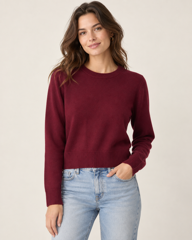 A woman wearing a burgundy knit sweater and light blue jeans, photographed from the back against a plain light beige studio background.