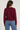 A woman wearing a burgundy knit sweater and light blue jeans, photographed from the back against a plain light beige studio background.
