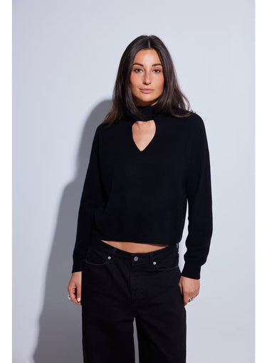 A woman with long dark brown hair wearing a black knit mock neck sweater with a heart-shaped keyhole cutout, photographed against a light gray studio background.