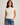 A woman wearing a cream-colored short-sleeve knit top with a button-down front and a high mock neck, paired with blue jeans, against a plain light gray background.
