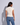 A woman shown from the back wearing a fitted short-sleeve cream knit top and blue jeans against a light gray studio background.