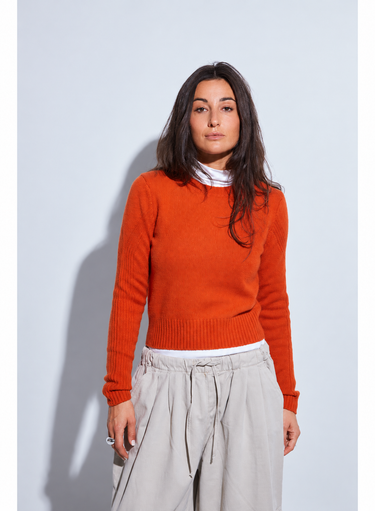 Woman wearing a bright orange crewneck sweater layered over a white mock-neck top with relaxed light beige wide-leg pants.