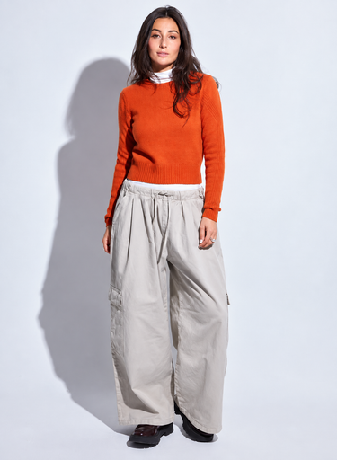 Woman wearing a bright orange crewneck sweater layered over a white mock-neck top with relaxed light beige wide-leg pants.