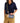 A person wearing a navy shirt and khaki pants holds a blue striped clutch bag with a colorful striped strap and a small gold-tone keychain. The bag has a tan leather brand patch that reads 'CONSUELA.