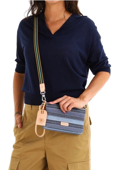 A person wearing a navy shirt and khaki pants holds a blue striped clutch bag with a colorful striped strap and a small gold-tone keychain. The bag has a tan leather brand patch that reads 'CONSUELA.