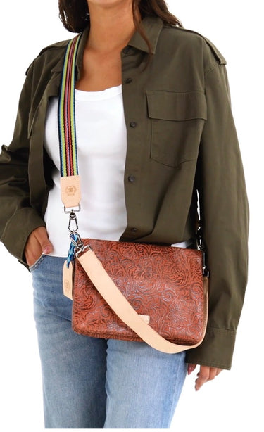 A woman wearing an olive green shirt and blue jeans carries a brown embossed leather crossbody bag with a beige strap and a colorful striped shoulder strap detail.