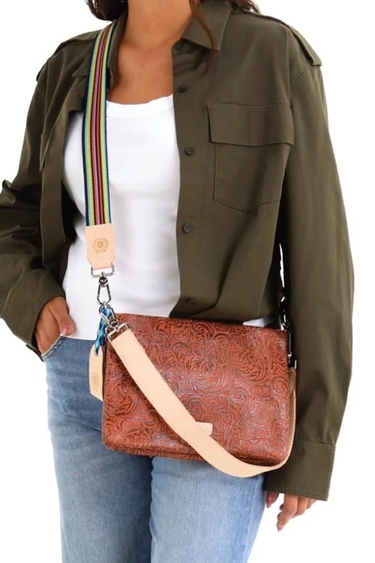A woman wearing an olive green shirt and blue jeans carries a brown embossed leather crossbody bag with a beige strap and a colorful striped shoulder strap detail.
