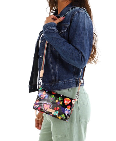 A woman wearing a Consuela black crossbody bag with colorful heart and floral print, tan wristlet strap, and striped woven shoulder strap.