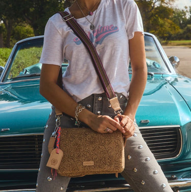A person wearing a white graphic t-shirt and gray studded jeans sits in front of a teal vintage convertible, carrying a brown embossed crossbody bag with a striped strap.