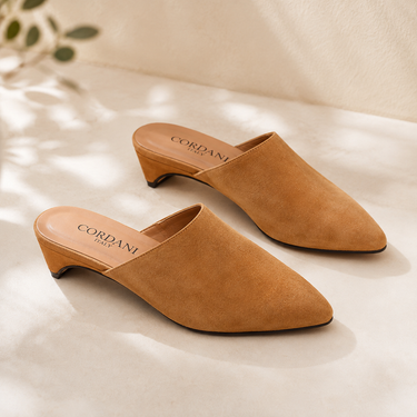 A pair of tan suede mule heels with low block kicks, shown on a light beige textured surface with soft leaf shadows. The brand name CORDANI is printed on the insole.