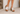A person wearing a light beige dress and metallic silver open-toe mule sandals with chunky brown wooden block heels, standing against a textured off-white wall.