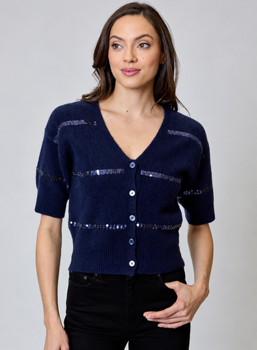 A woman wearing a navy short sleeve v-neck cardigan with blue sequin stripes and black jeans on a neutral background