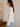 Woman wearing a white sweater and blue jeans standing in a room with a vase and painting on a wall.