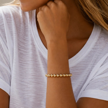 A woman wearing a gold beaded stretch bracelet made of polished metallic spherical beads.