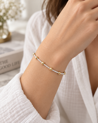 A delicate gold and silver beaded bracelet worn on a woman's wrist, styled with a white textured robe in a bright, softly blurred indoor setting.