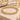 A gold beaded bracelet made of polished metallic round beads, arranged in a circle on a light beige stone surface with soft decorative props in the background.