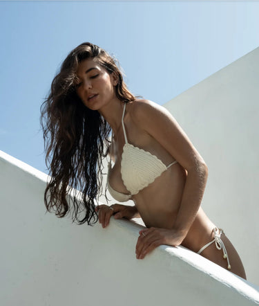 Woman in a white crochet bikini leaning against a white wall with a blue sky background