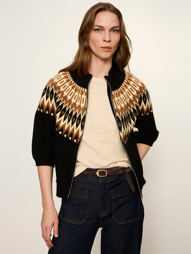 A woman modeling a black knit zip-up jacket with a tan and beige geometric pattern across the shoulders and upper chest, paired with a cream t-shirt and dark blue jeans against a plain light background.
