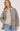 A woman wearing a quilted taupe-gray jacket with a beige collar, layered over a beige knit sweater and blue jeans, against a light gray studio background.
