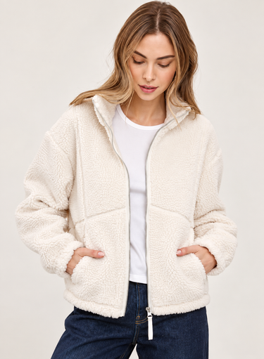 A woman wearing a cream sherpa fleece zip-up jacket over a white t-shirt and dark blue jeans, photographed against a neutral light gray studio background.