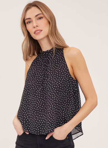 A woman wearing a sleeveless black and white polka dot flowy blouse with a mock neck and black jeans on a white background.