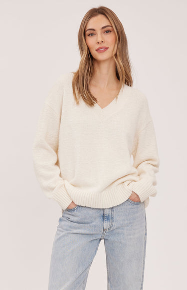A woman wearing a cream v-neck pullover and blue jeans on a white background