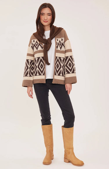 A woman modeling a brown and beige patterned knit cardigan with a matching brown scarf, paired with black pants and tan boots, against a light beige studio background.