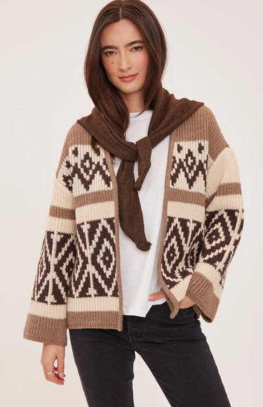 A woman wearing a brown, cream, and dark brown knit sweater with geometric patterns, shown from a three-quarters back view against a light beige studio background.