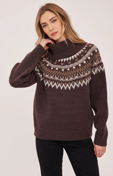 A woman wearing a brown turtle neck with a fair isle print and black jeans on a white background.