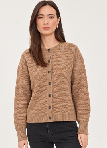 A woman wearing a cropped, long-sleeve knit cardigan in a warm camel brown color with dark buttons, paired with black jeans, against a plain light background.