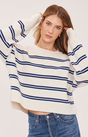 A woman wearing a cream and navy stripe oversized sweater and blue jeans on a white background